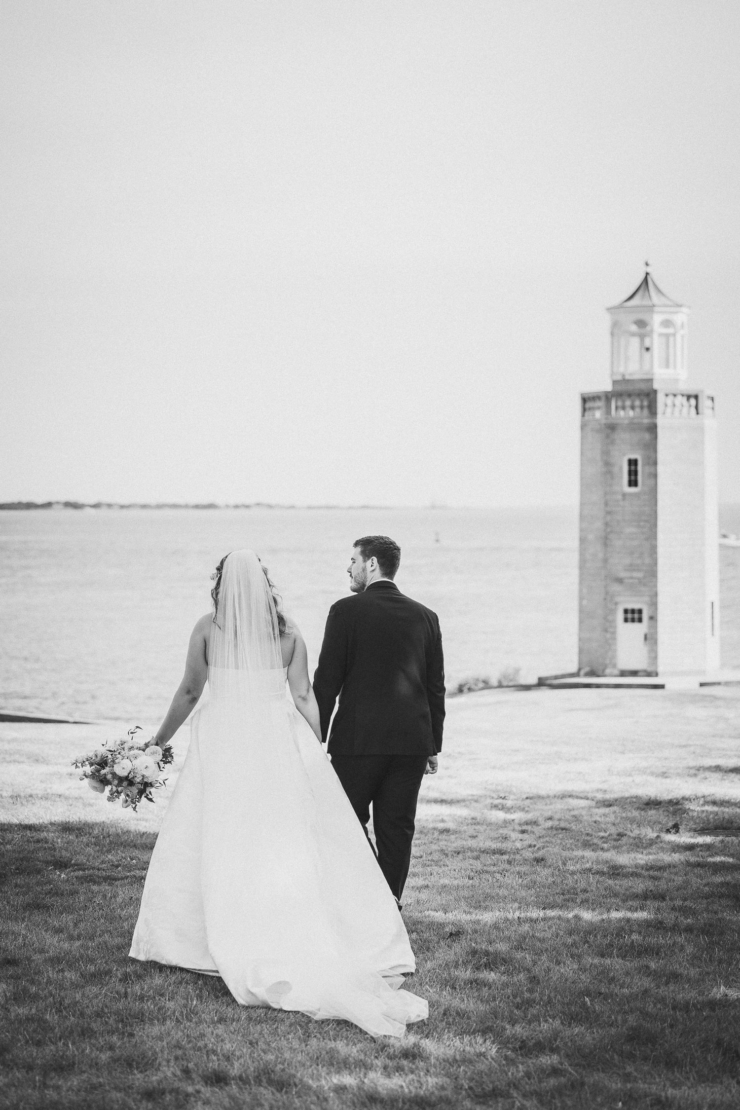 A romantic black and white wedding photo series showing a couple walking towards a lighthouse on a grassy coastline.