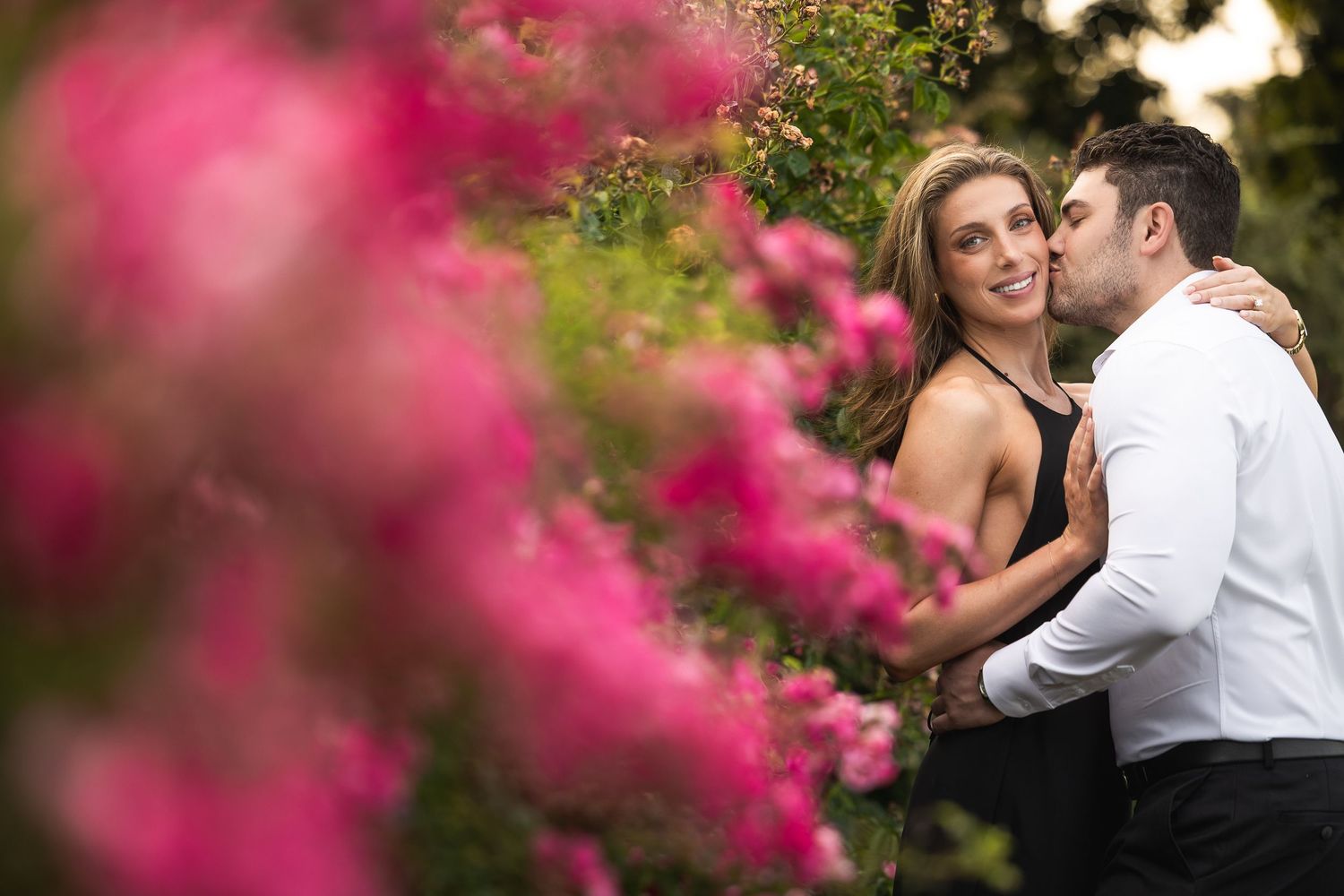 A romantic couple embraces in a garden with vibrant pink flowers creating a dreamy foreground blur.
