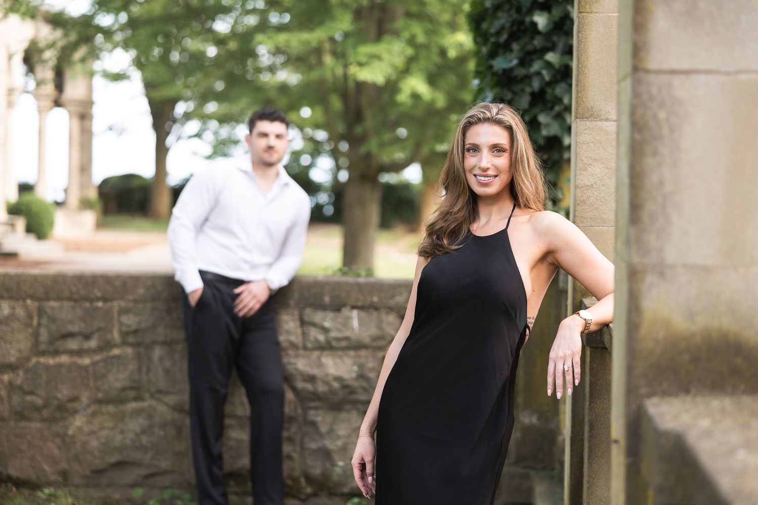 A woman in a black dress poses by a stone wall while a man in white stands in the background.