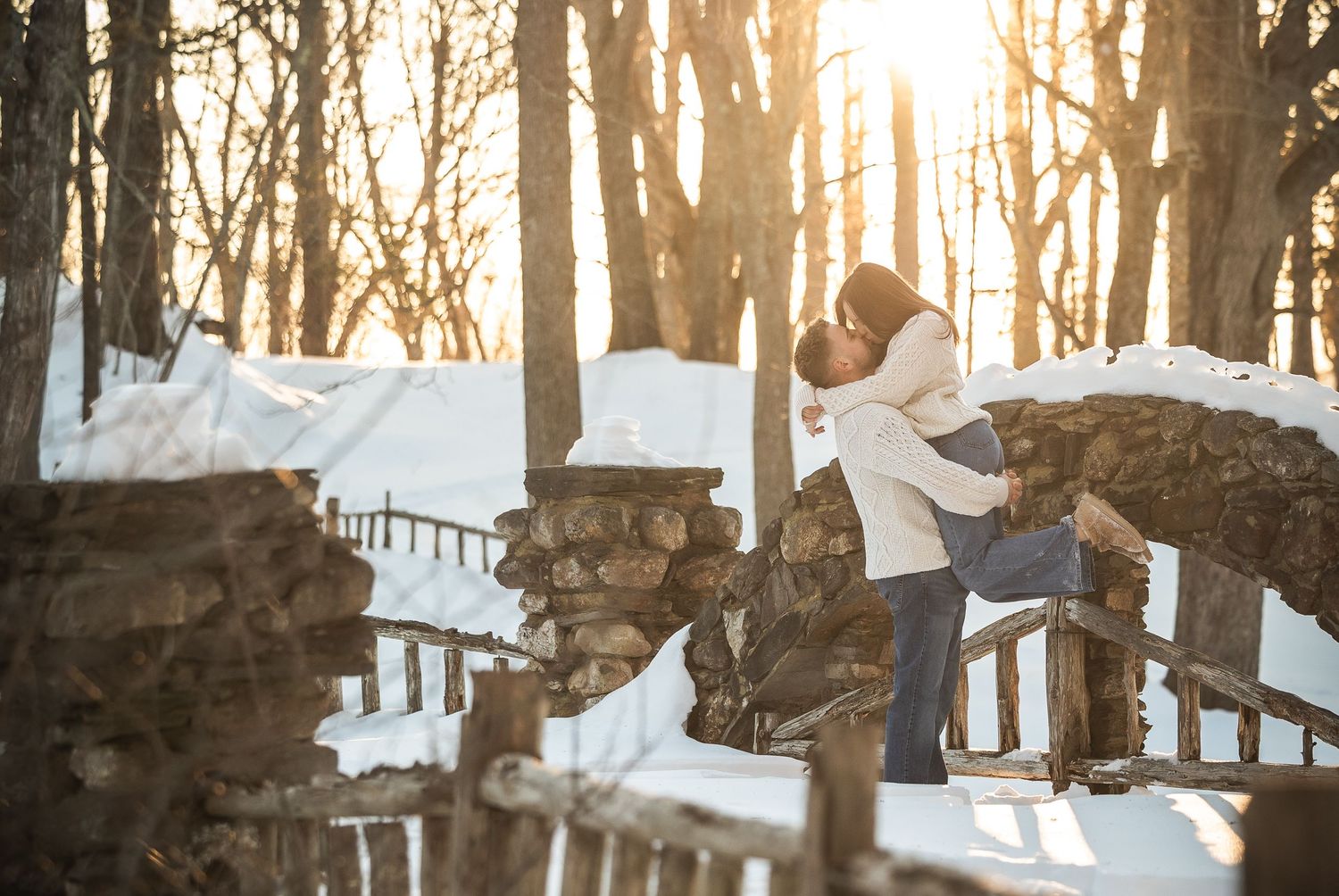 Snowy winter engagement photo session in a wooded area with warm golden sunlight filtering through bare trees.