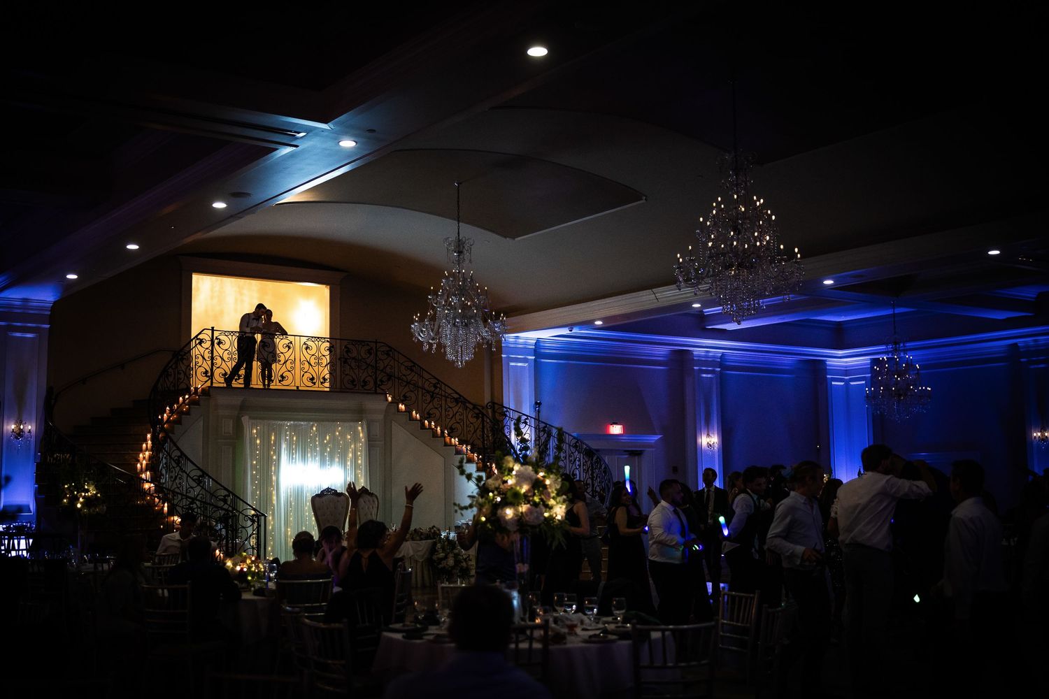A wedding reception venue decorated with blue uplighting and crystal chandeliers creates an elegant evening atmosphere.