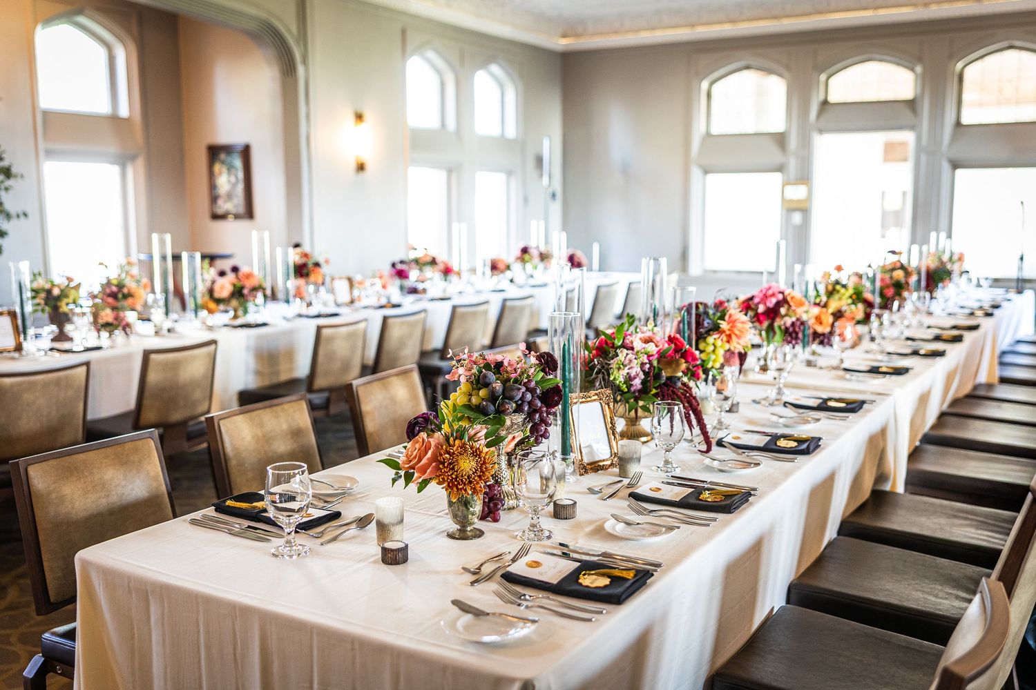 Long banquet tables with elegant place settings and colorful floral centerpieces arranged in a bright, sunlit dining room.