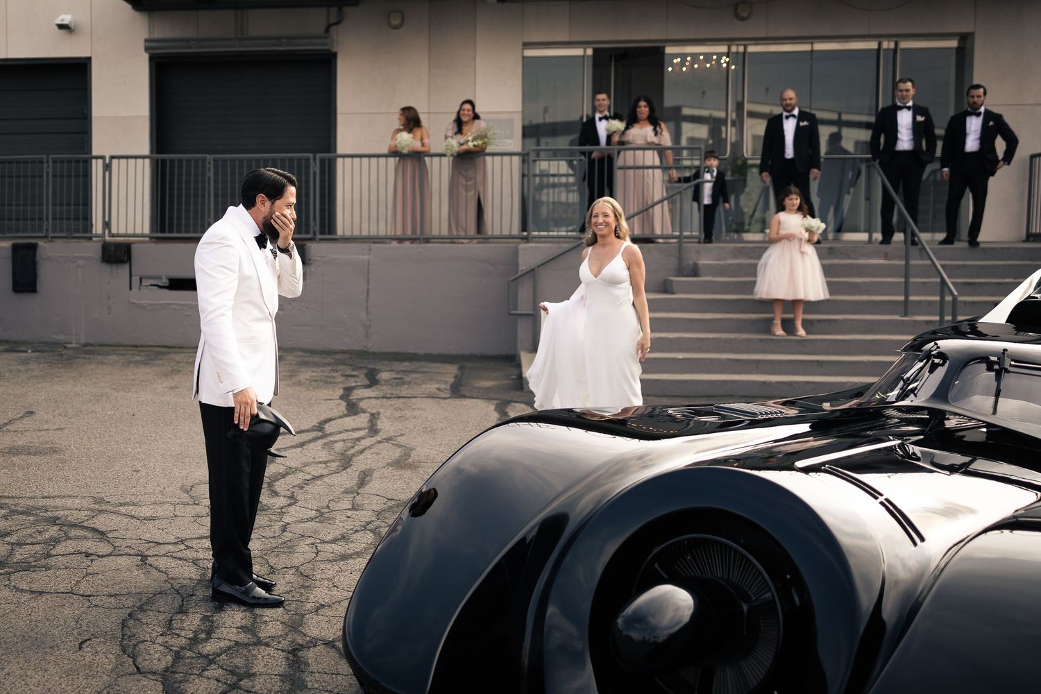 A vintage Batmobile replica parked at a wedding venue with people gathered on the steps.