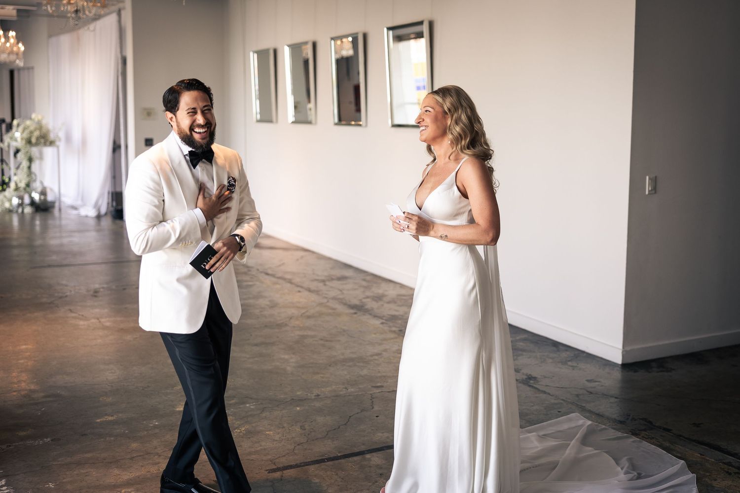 A couple in wedding attire playfully interacts in a bright industrial space with white walls and concrete floors.