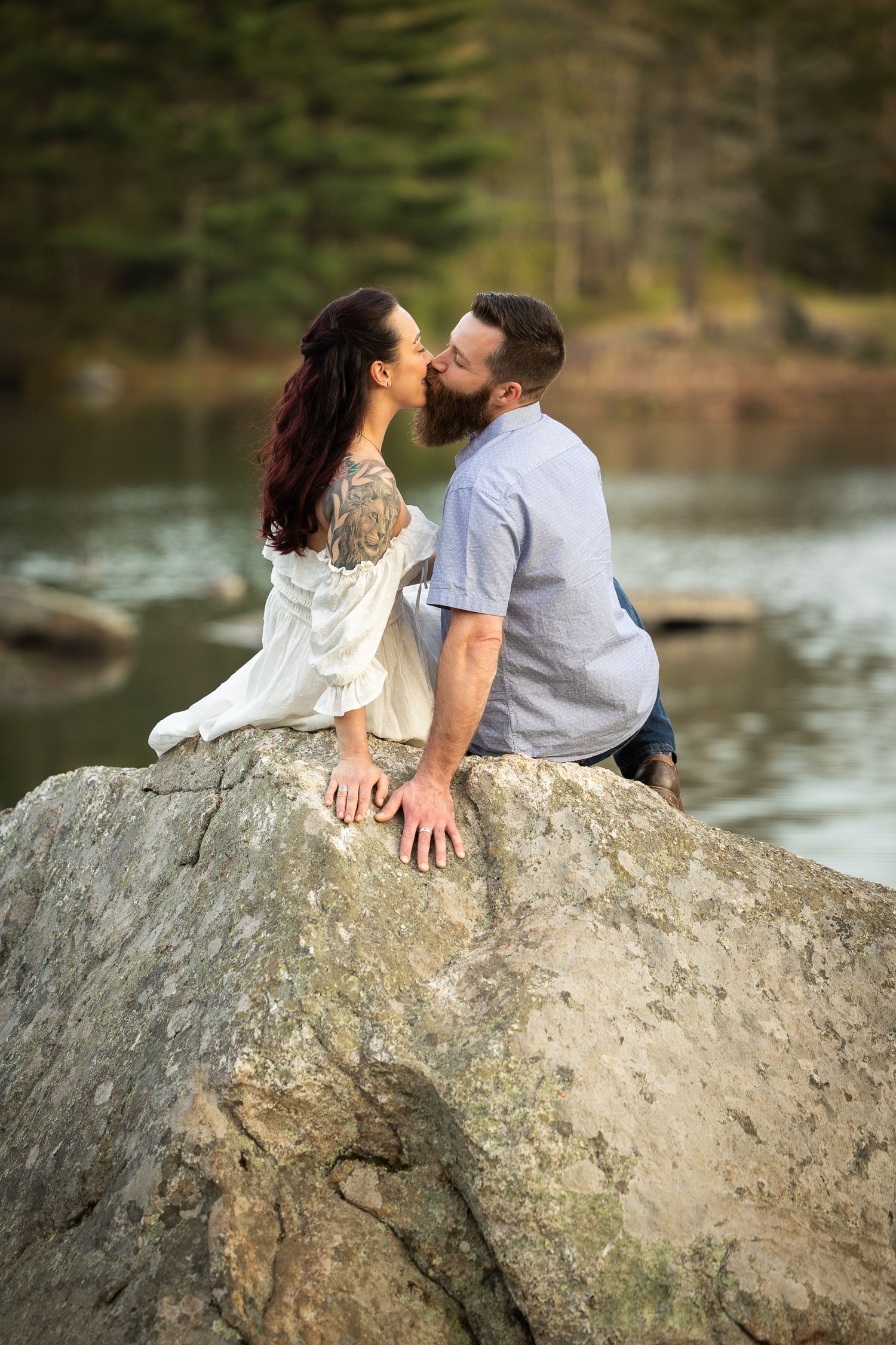 A romantic couple shares a tender kiss while sitting on a large boulder beside a serene lake in a forest setting.