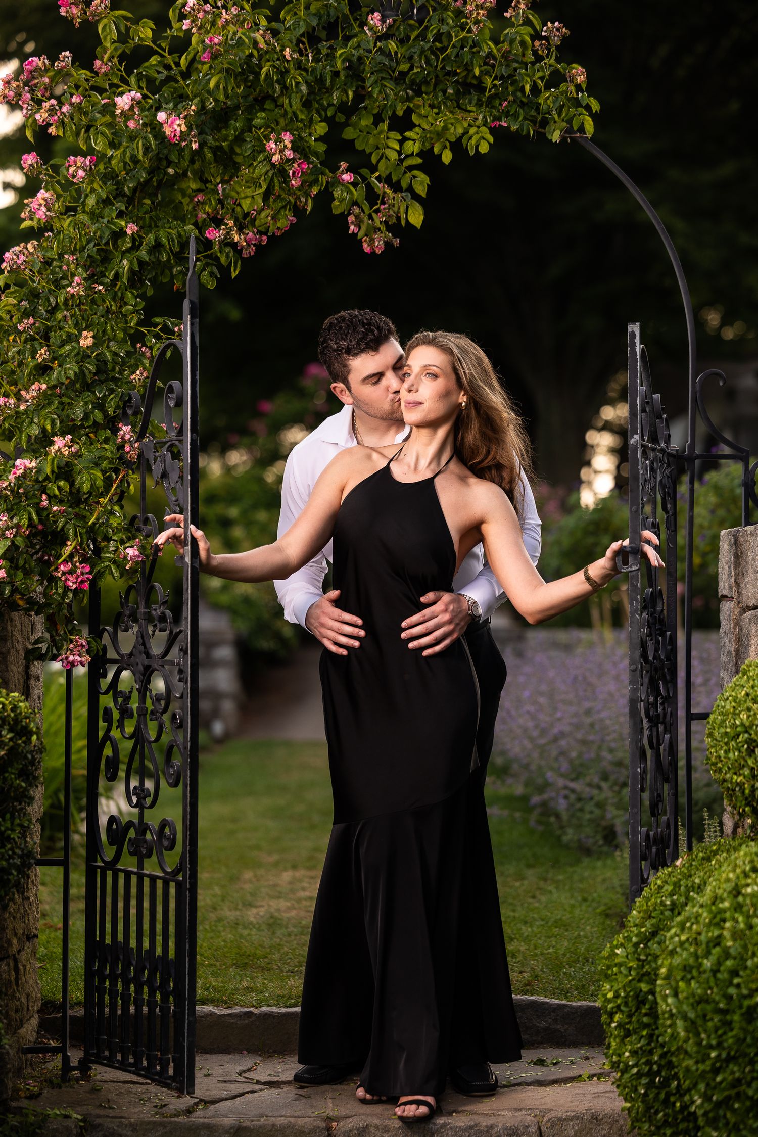 A romantic couple embraces near a garden gate surrounded by blooming pink flowers and lush greenery.