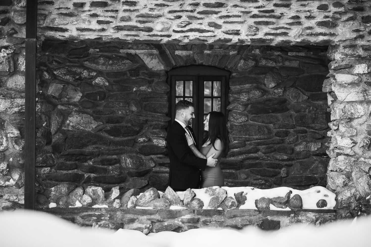 A black and white photograph of someone looking through a rustic stone cottage window with snow on the ledge.