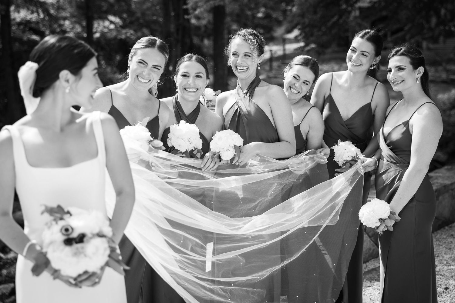 Black and white photo of bridesmaids holding a wedding veil in an outdoor setting.
