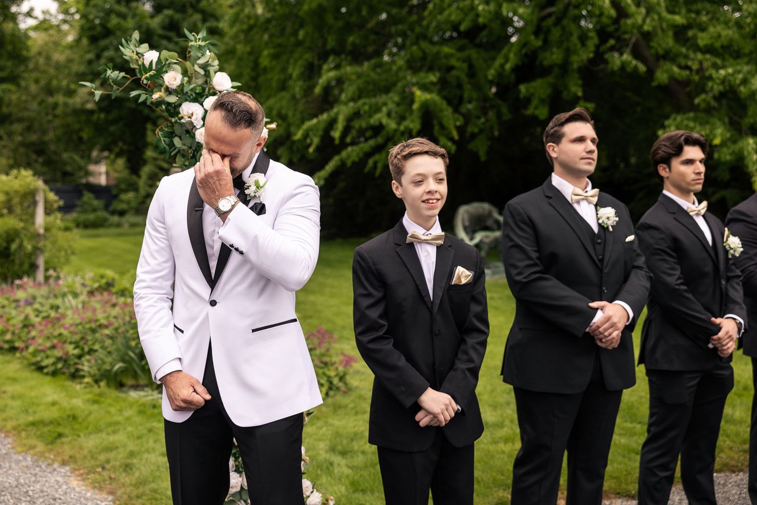 Groomsmen in black suits stand in a line outdoors against green foliage.