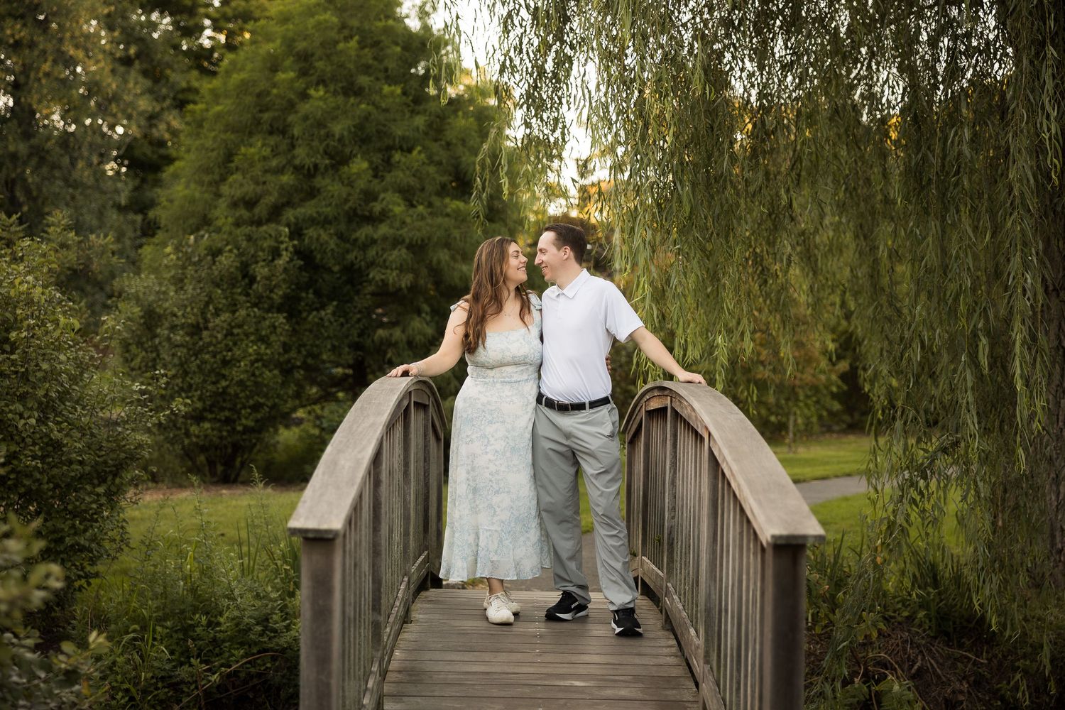 Couple sharing a romantic moment on a wooden bridge surrounded by lush green trees and weeping willows in a park.