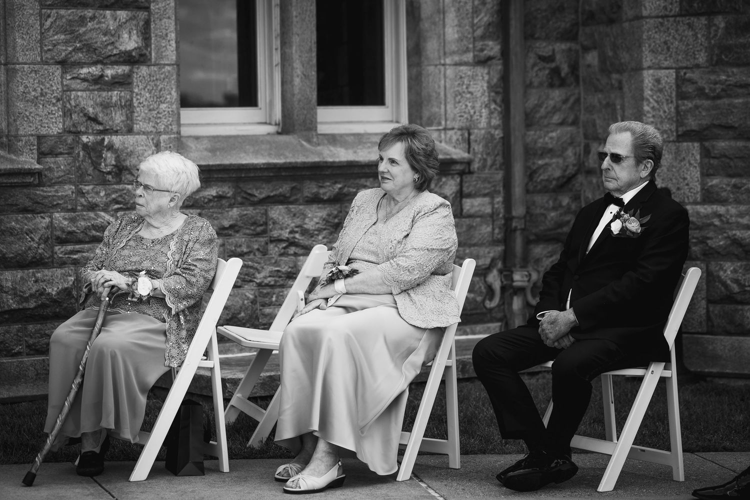 Elderly residents relax in white chairs along the stone wall of a care facility.