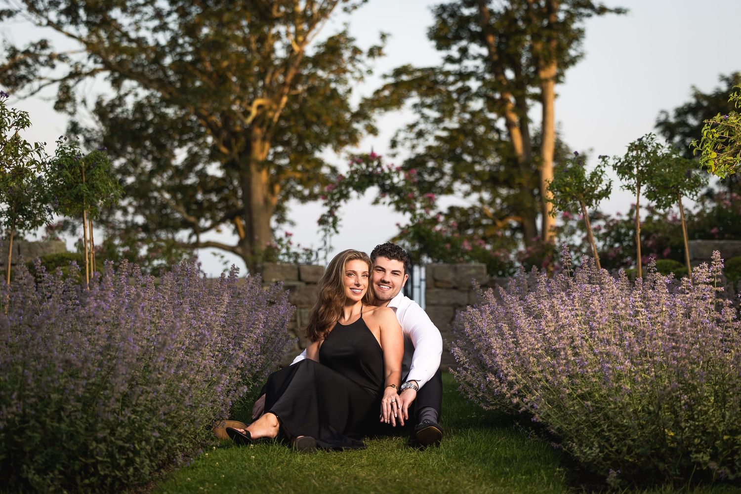Couple shares intimate moments in outdoor garden setting with purple flowers and trees at sunset.
