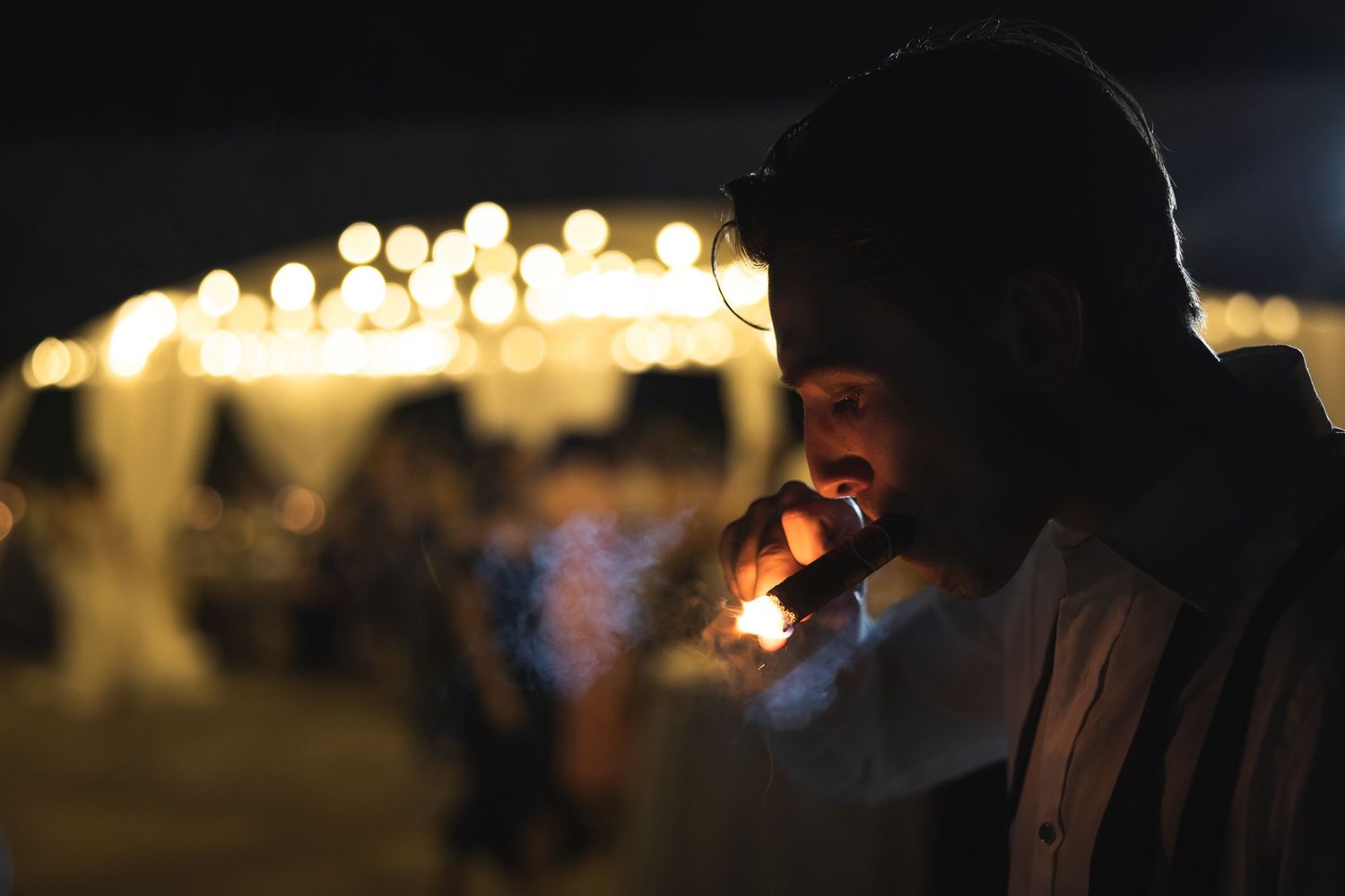 Groom lighting a cigar while an outdoor wedding reception takes place in the background.