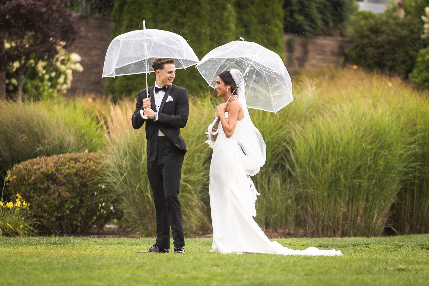 Wedding couple holding clear umbrellas while posing on lush garden grounds.