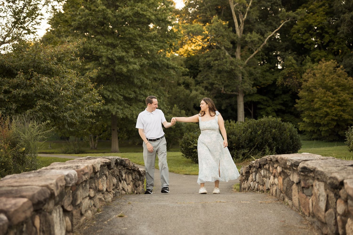 Couple holding hands walking on a stone bridge surrounded by lush green trees in a park setting.