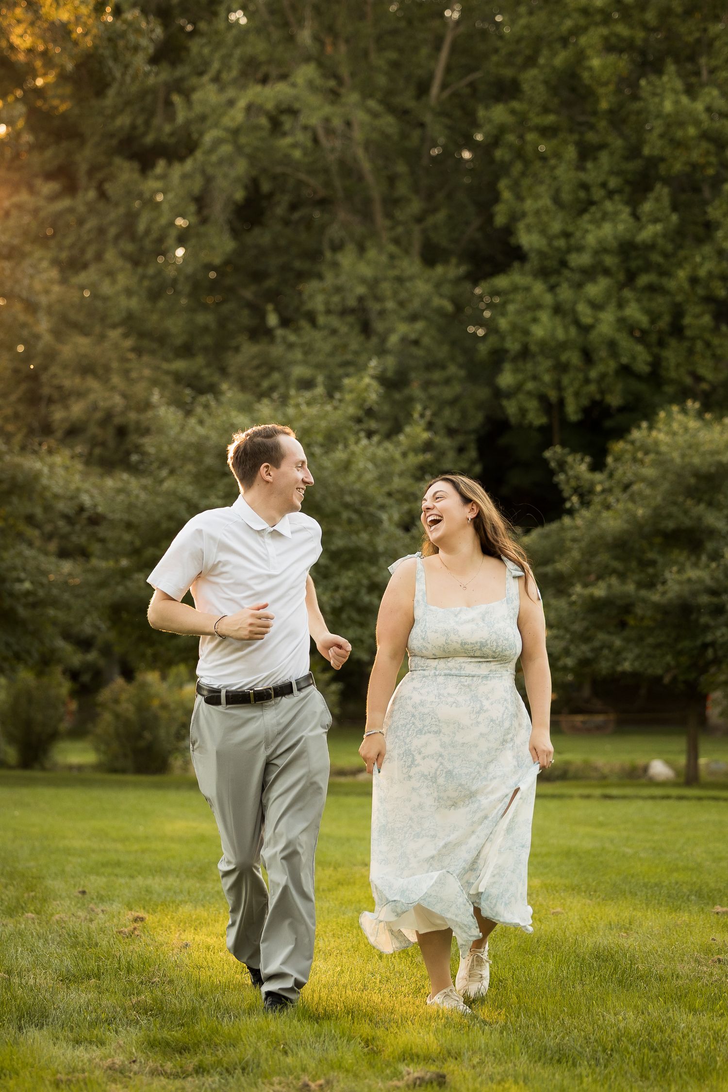 A laughing couple walks through a sunlit green park, the woman in a floral dress and the man in a white shirt and gray pants.