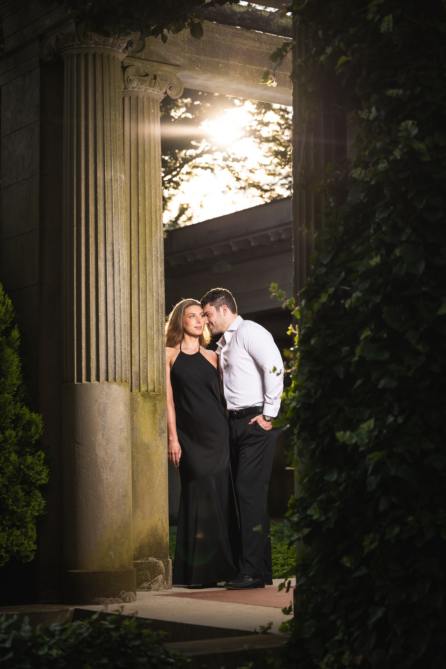 A romantic couple shares an intimate moment between stone columns at dusk in a garden setting.