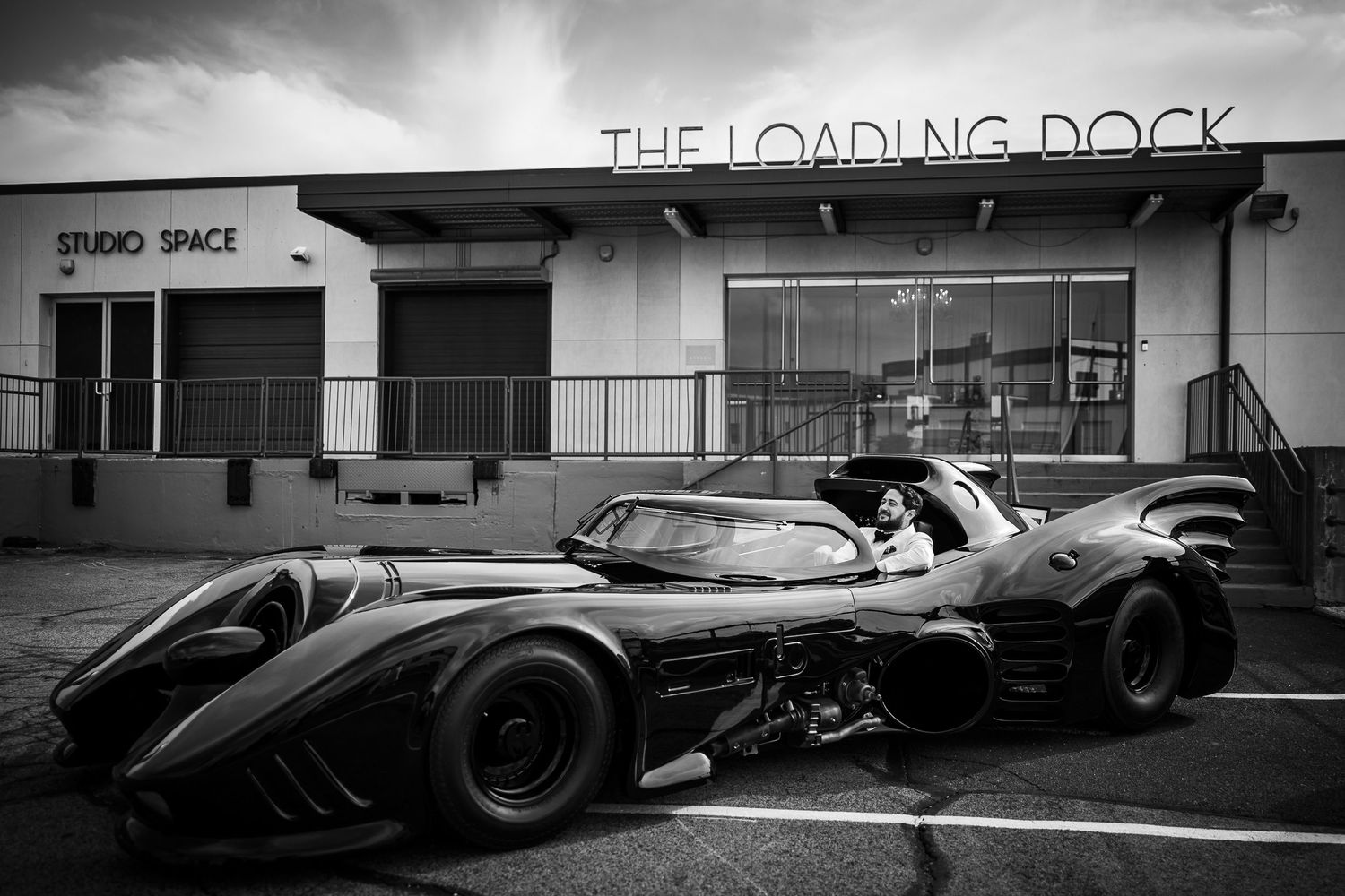 A black and white photograph shows a vintage race car on a track with moody cloudy skies in the background.