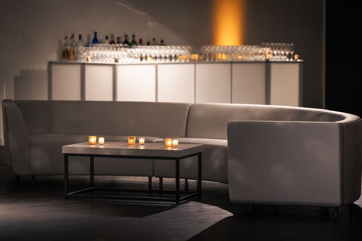 Modern lounge area with curved gray seating, coffee table with candles, and illuminated bar shelving in the background.