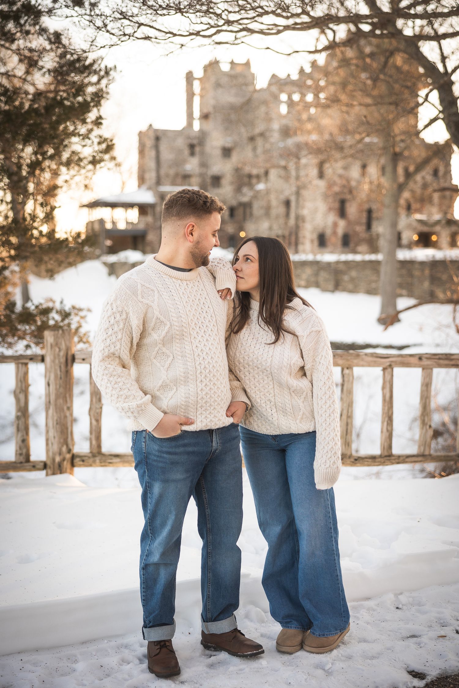 Couple in matching cream sweaters and jeans walking through snowy winter landscape at sunset.