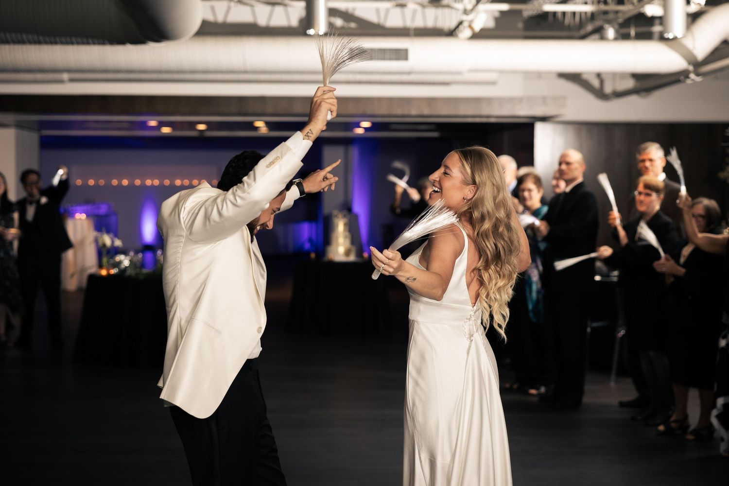 A couple in white formal attire dances energetically at a reception with a purple-lit backdrop and guests in the background.