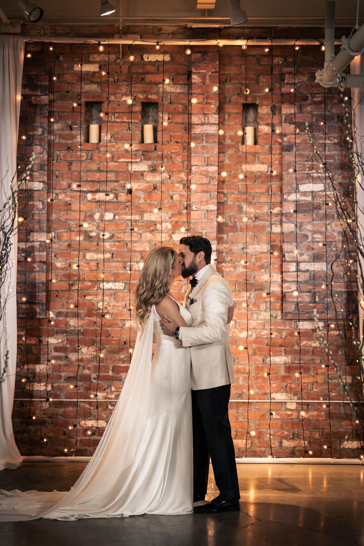 A romantic first dance moment captured against a rustic brick wall backdrop with twinkling string lights.