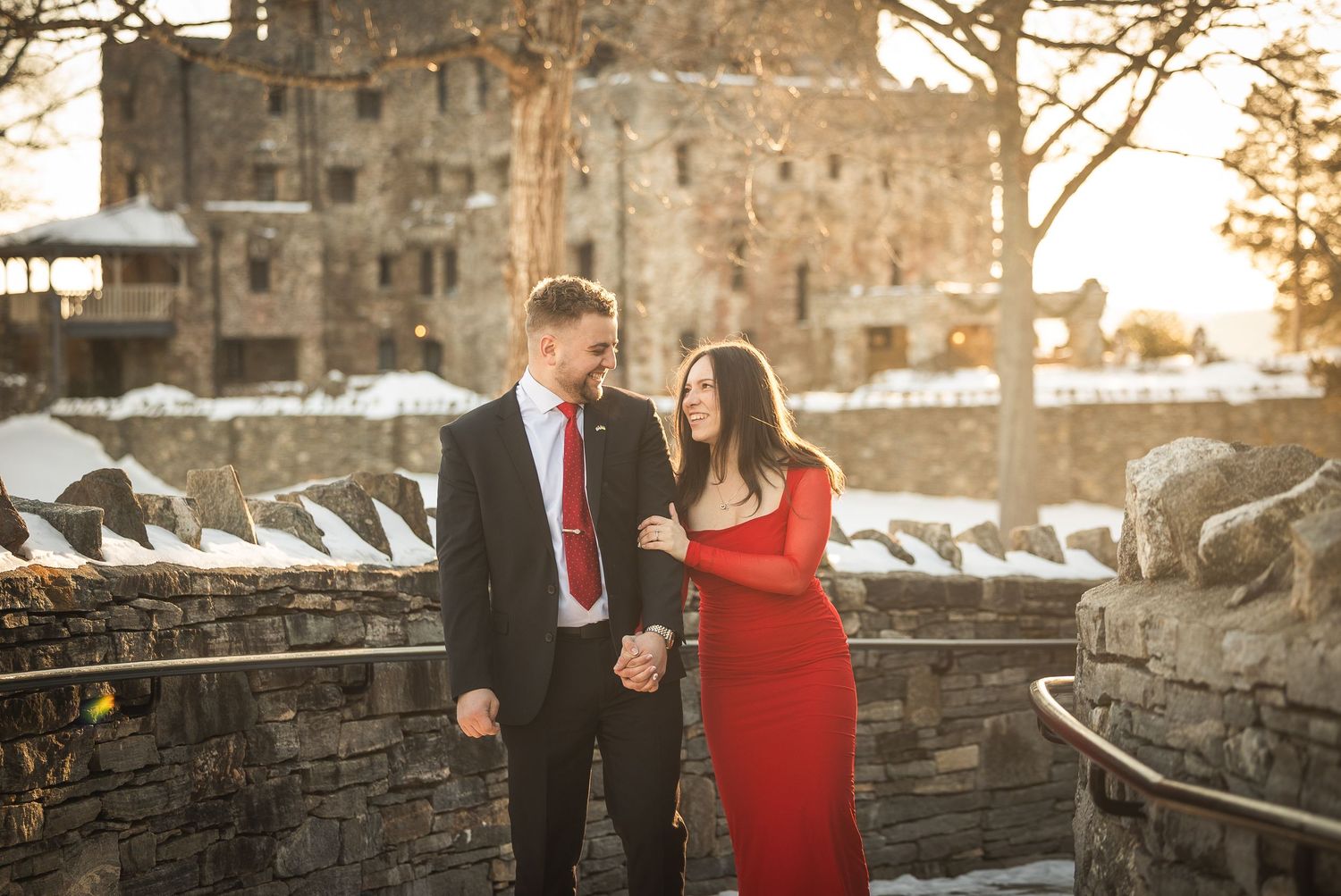 Couple in formal attire poses together on stone bridge during golden hour sunset.