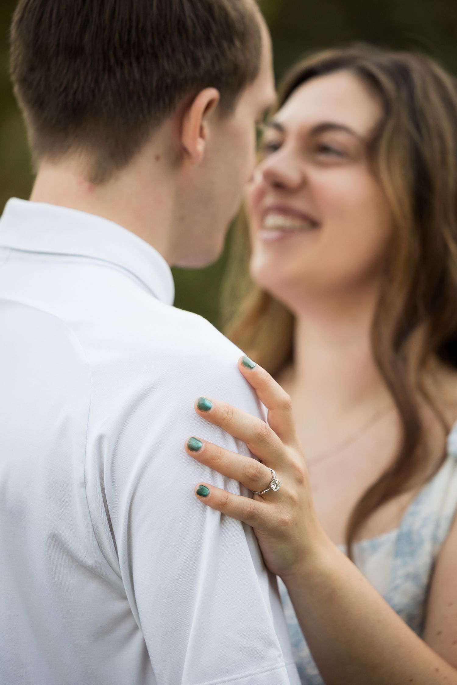 A smiling woman with teal nails and a ring rests her hand on a man's shoulder in a romantic outdoor portrait.