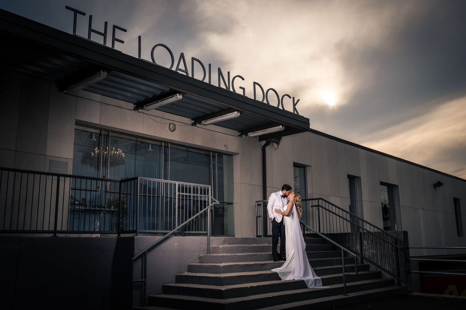 A dramatic wedding photo series showing a bride in a white dress against dark industrial architecture and moody skies.