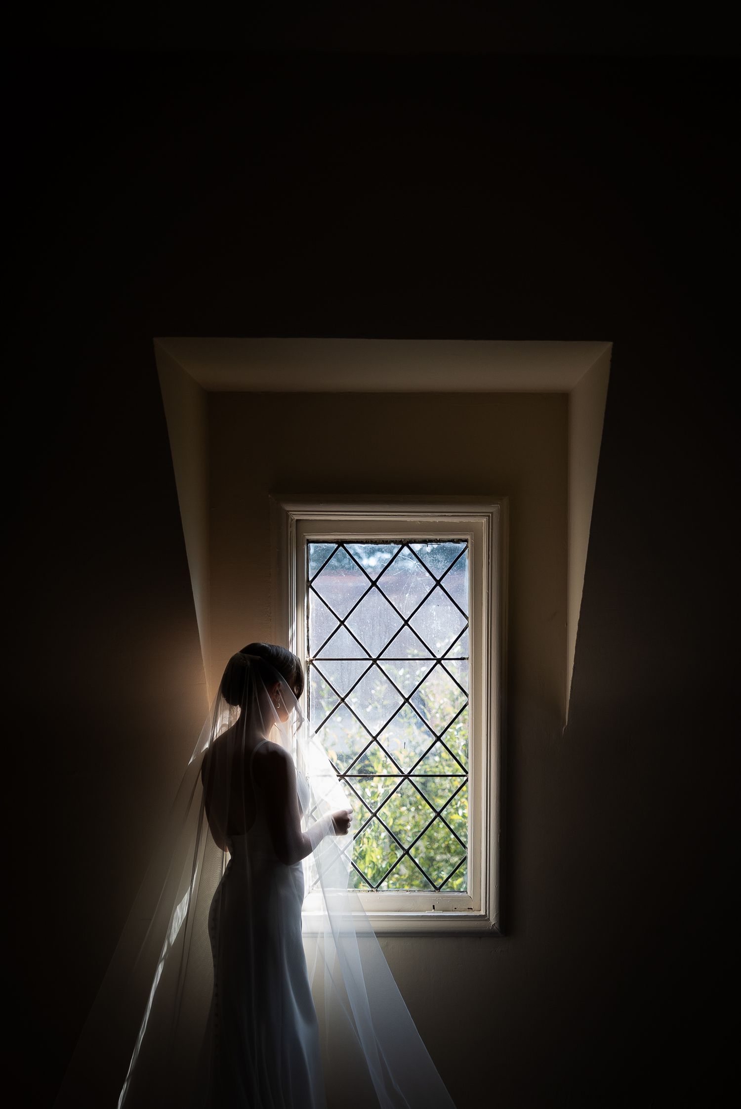 Silhouetted figure stands in dark room by diamond-paned window with dramatic natural light streaming through.