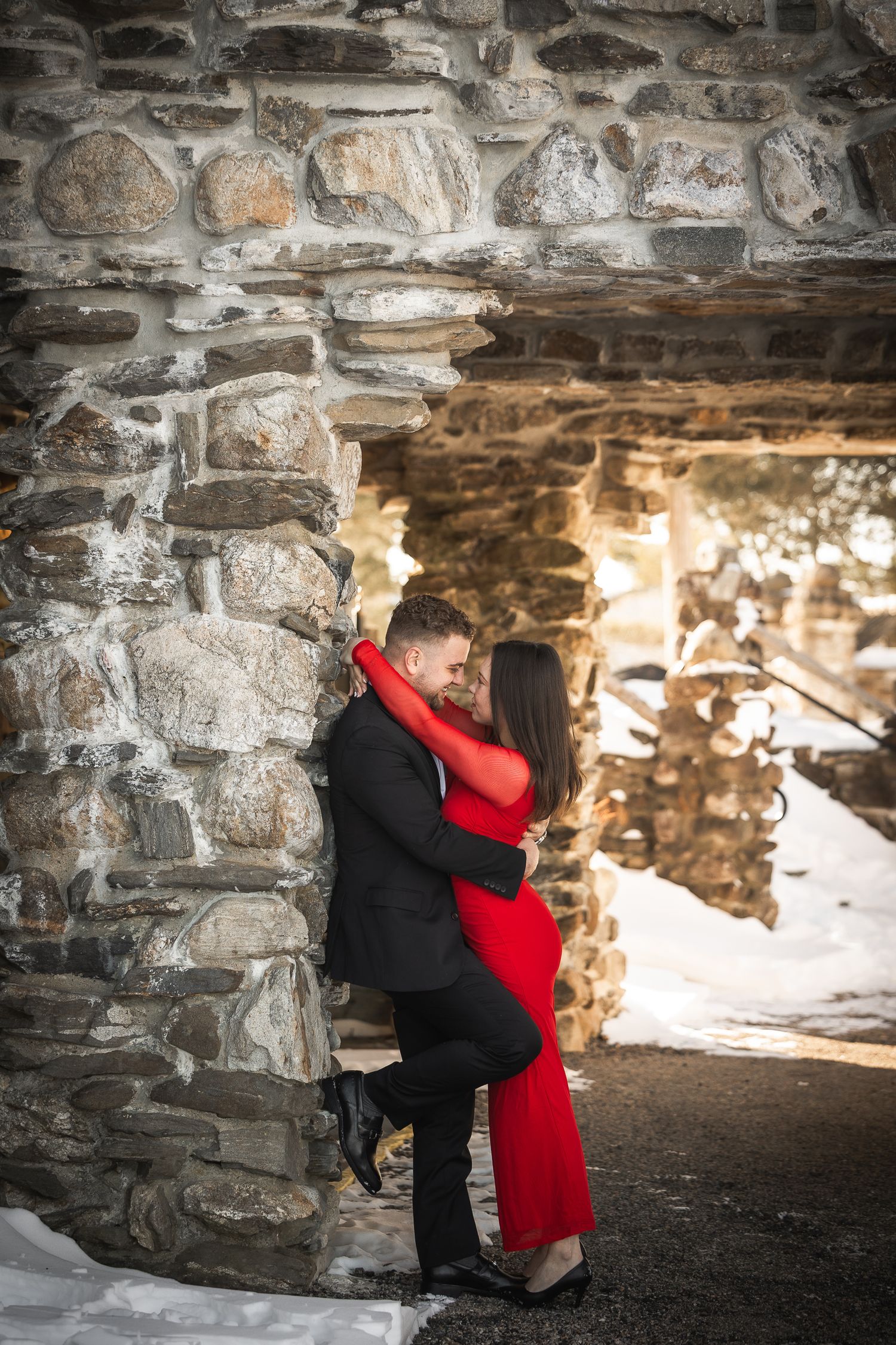 A couple in formal attire embraces under a stone archway during a romantic engagement photoshoot.