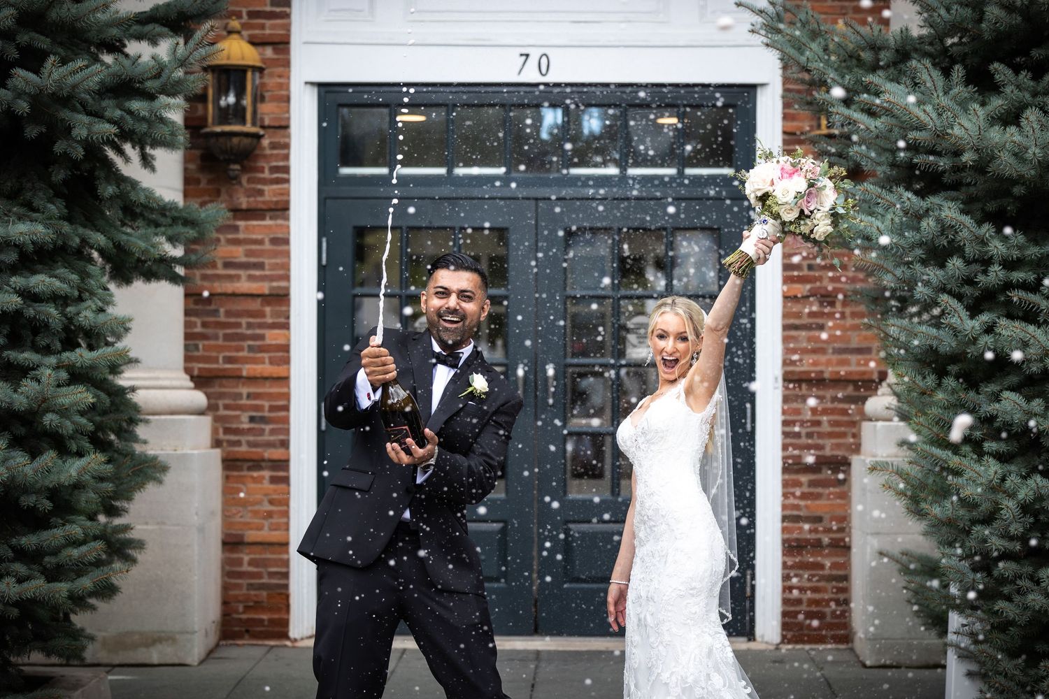 Wedding couple poses playfully against a rustic brick wall with evergreen trees.