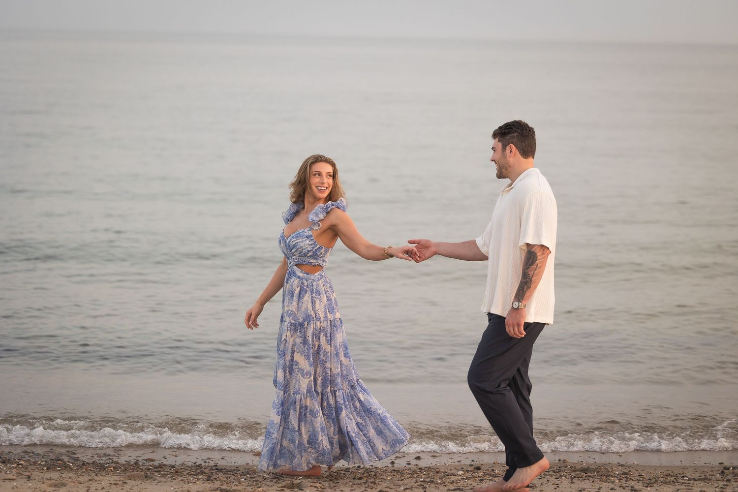 A couple walks along a misty beach at sunset while holding hands and wearing casual summer attire.