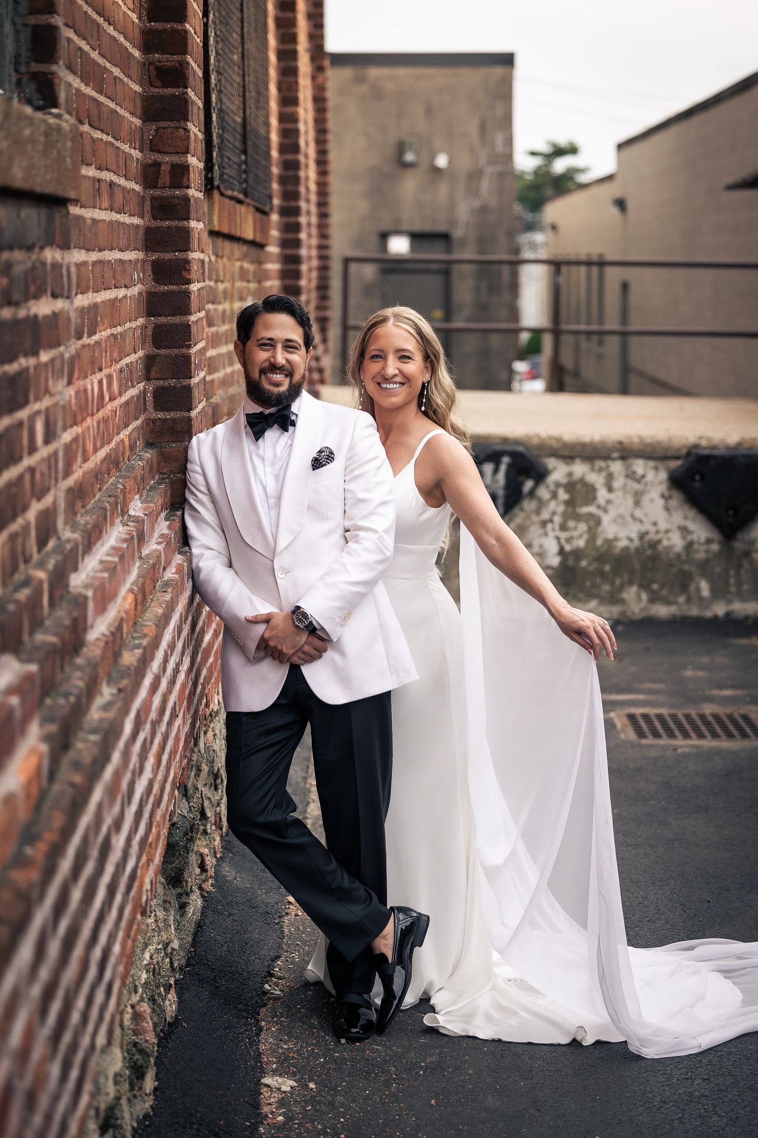 A couple in wedding attire poses together against a rustic brick wall backdrop in an urban setting.