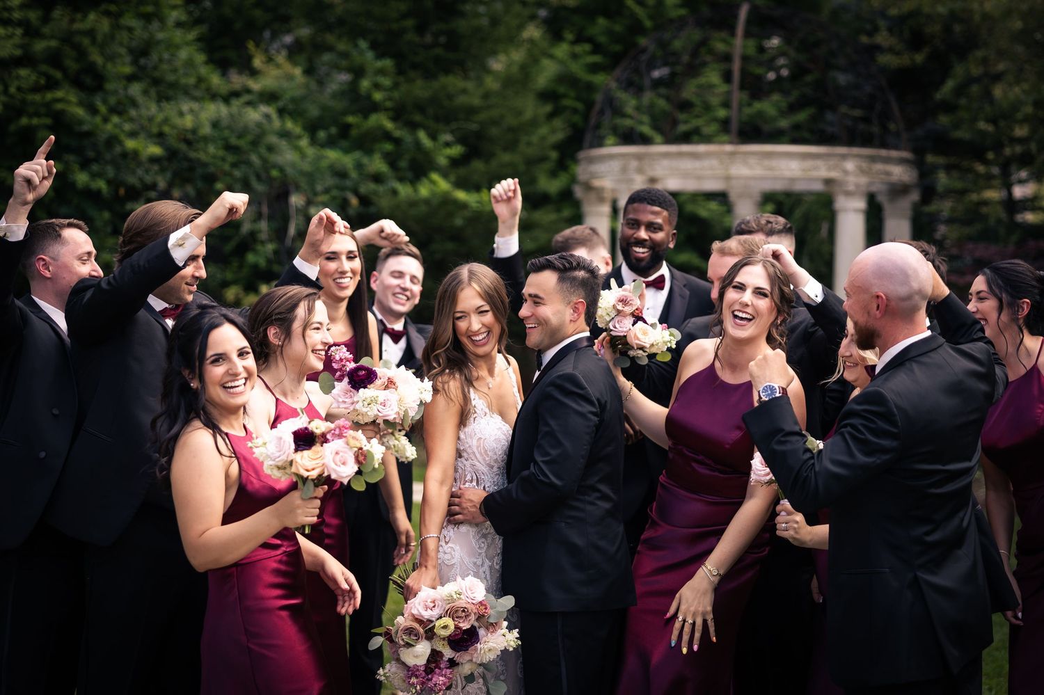 Wedding party celebrates joyfully outdoors with burgundy dresses and festive bouquets.