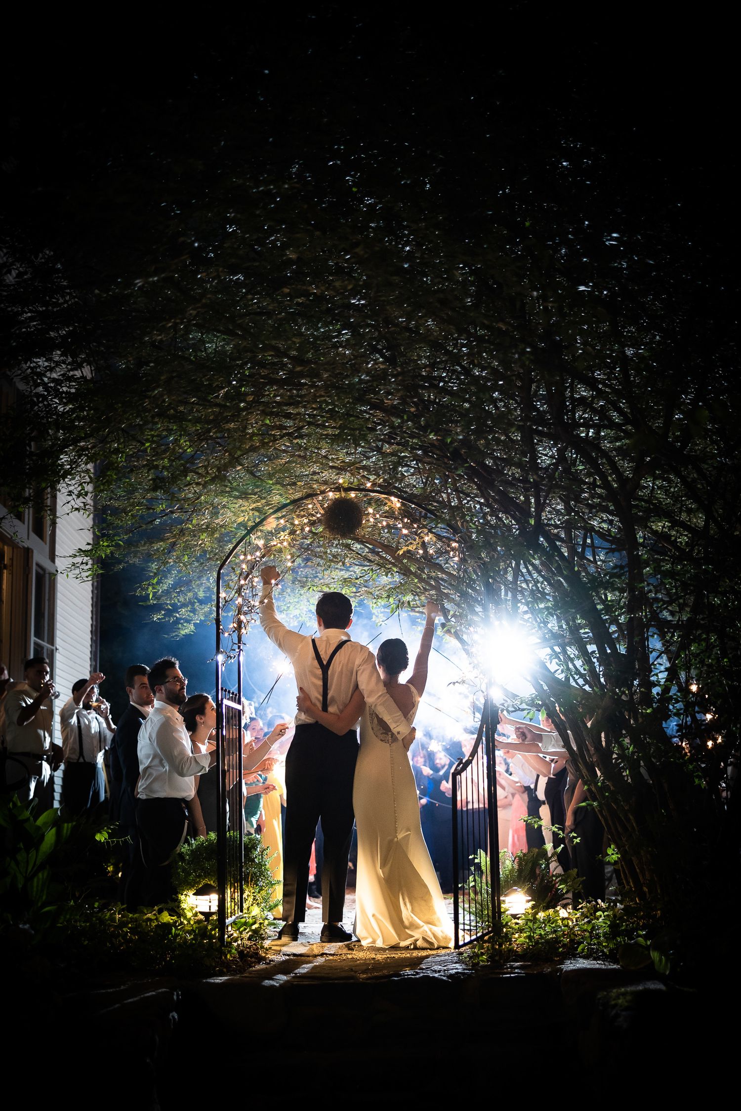 Silhouetted wedding couple exits through an illuminated garden archway at night.