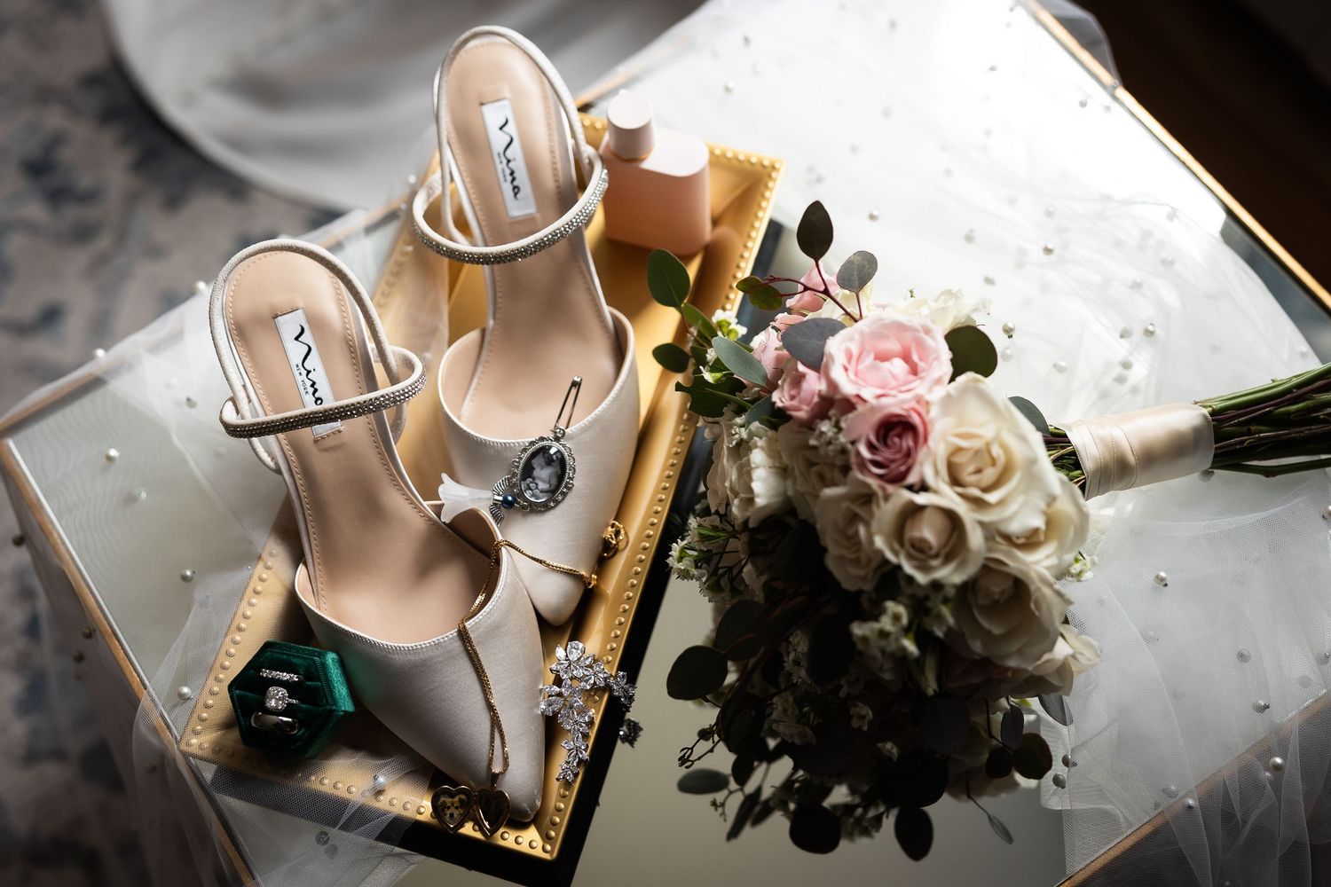 Elegant bridal accessories including beige heels and pastel floral bouquet on reflective surface.