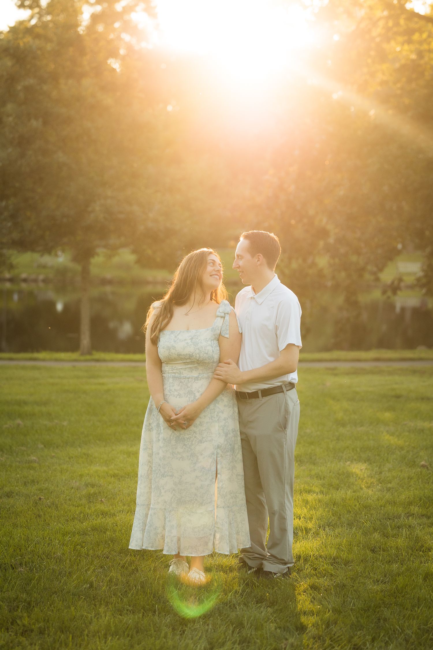 Couple embracing on green grass at golden hour sunset, woman in floral dress and man in white shirt, warm sunlight glowing behind them.