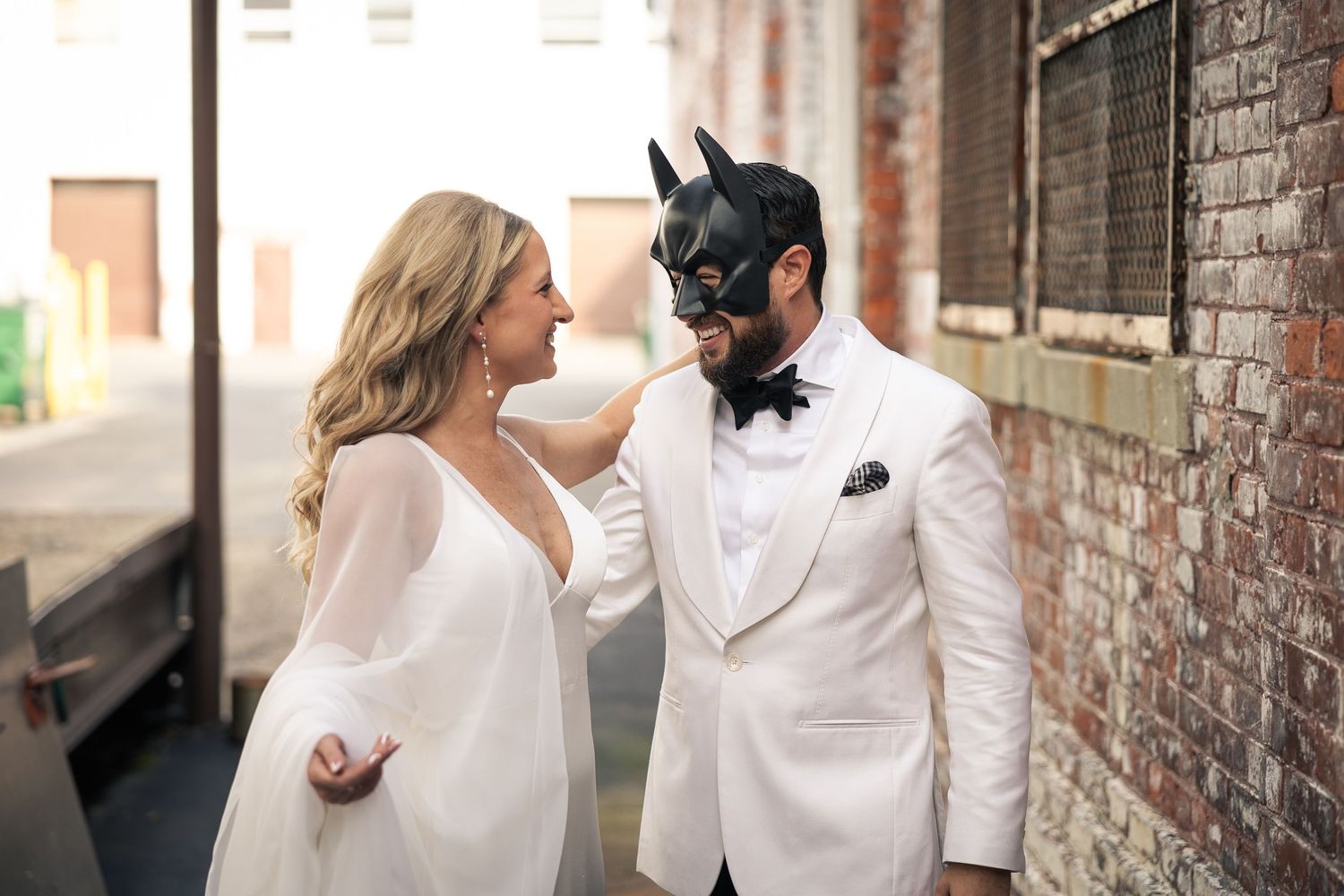 A couple in elegant white wedding attire share a romantic moment against a rustic brick wall backdrop.