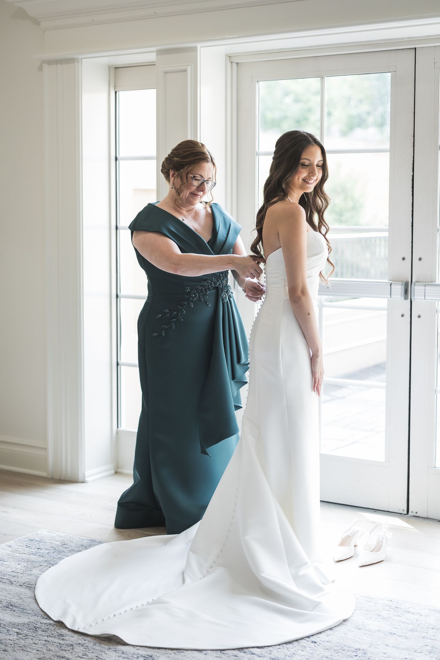 Helper assists with adjusting wedding dress train in bright white room with windows.