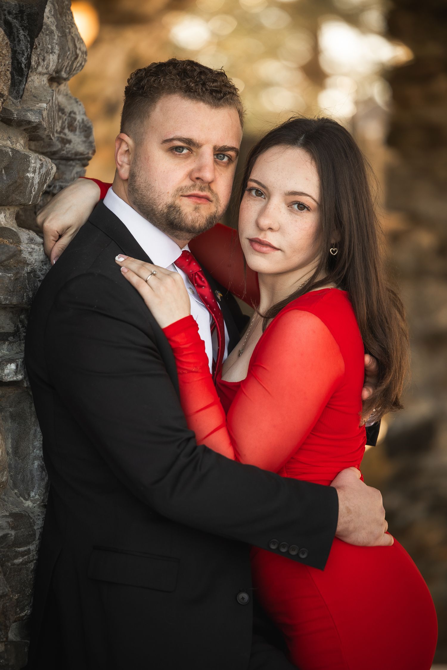 A romantic couple embraces against a stone wall backdrop, with her in a vibrant red dress and him in a black suit.