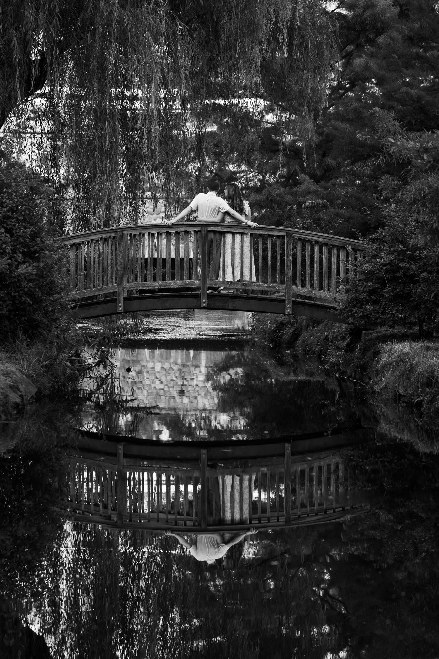 Couple standing on wooden bridge over reflective pond surrounded by weeping willows, black and white romantic garden photo.