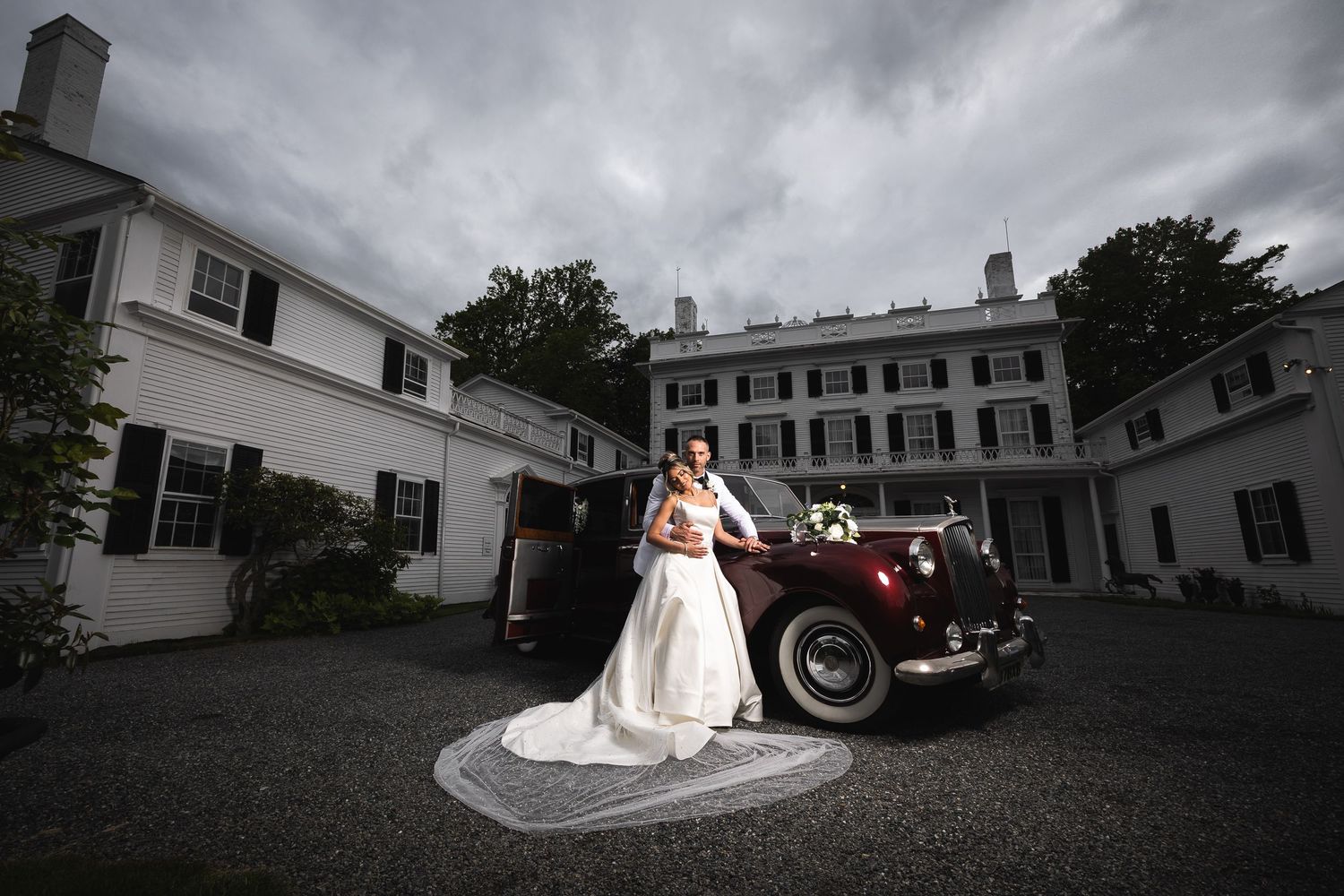 Dramatic wide-angle shot of wedding couple with vintage car under stormy skies.