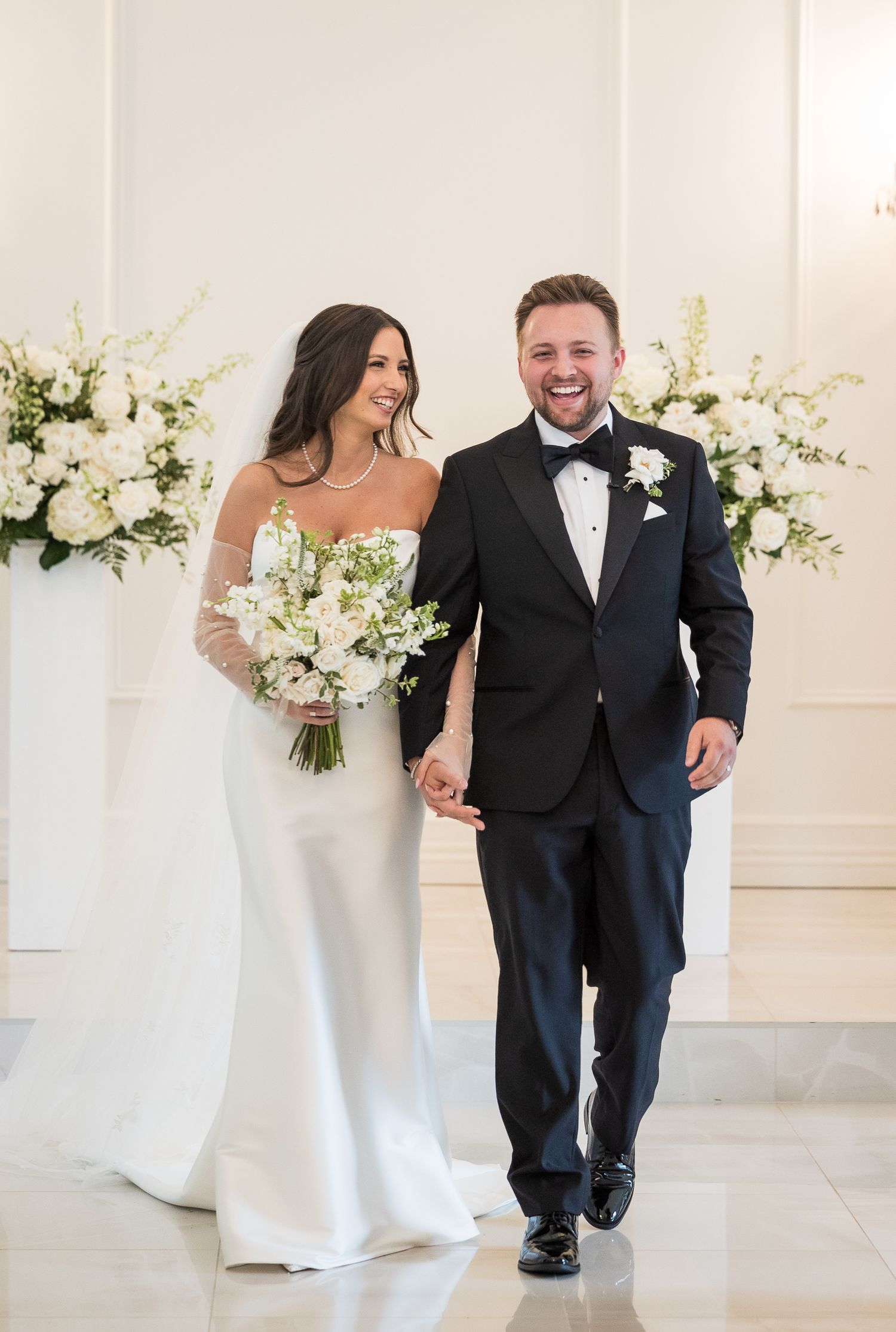 Wedding couple walks and laughs together in formal attire through an elegant white reception venue.
