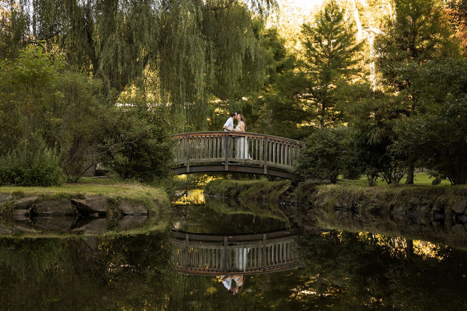 Couple embracing on a wooden bridge over a reflective pond surrounded by lush green trees in a serene garden.