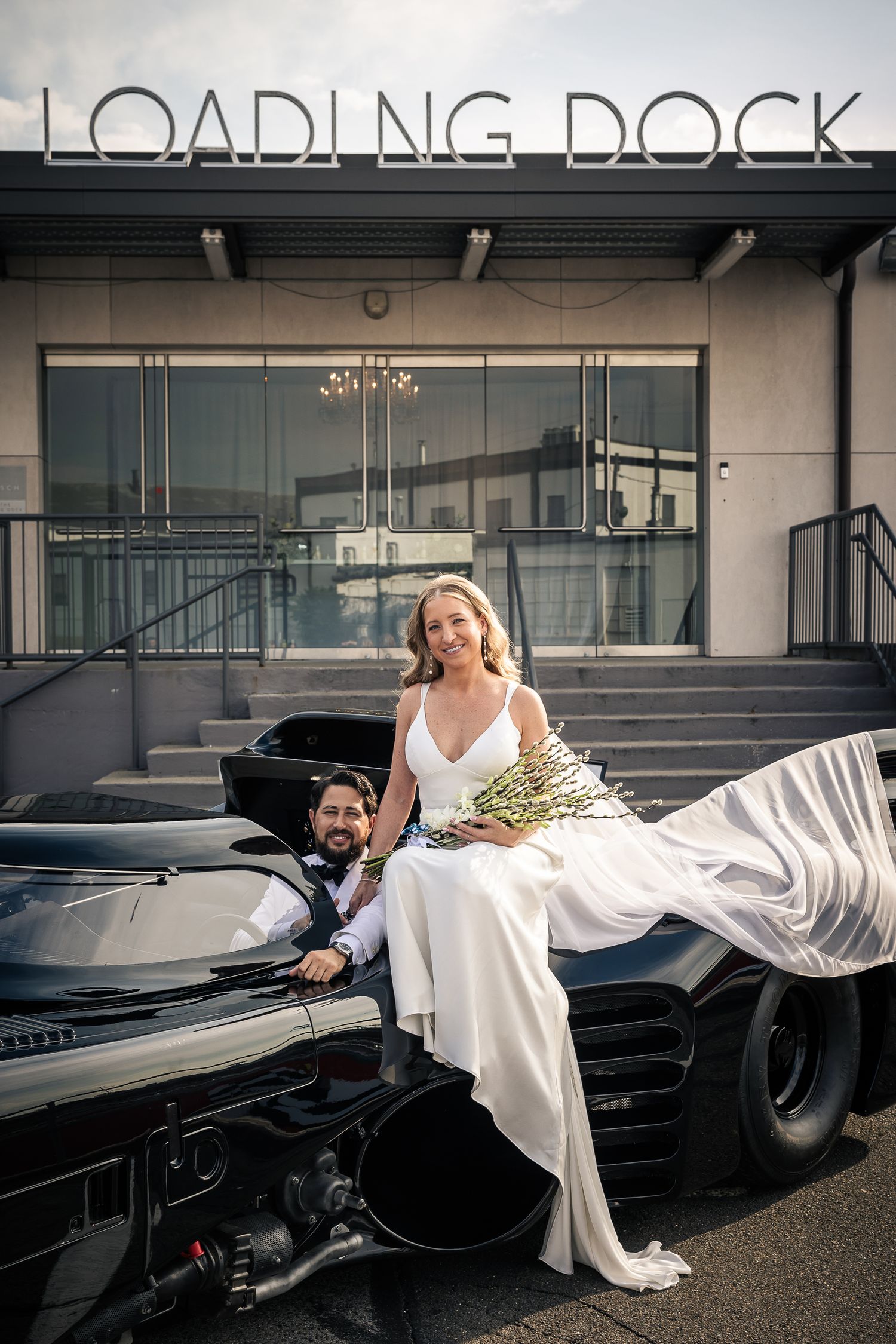 Wedding photography series at a loading dock featuring a white bridal gown draped over classic black cars.