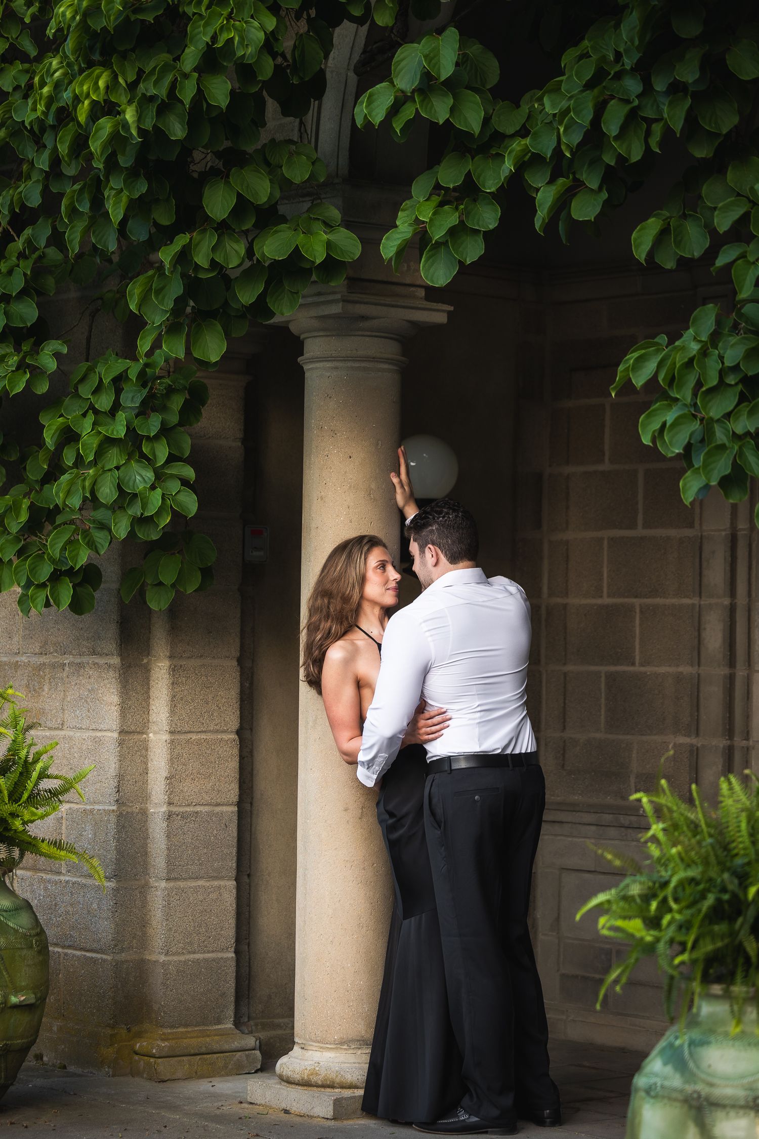 A romantic couple embraces in a stone archway surrounded by lush green foliage.