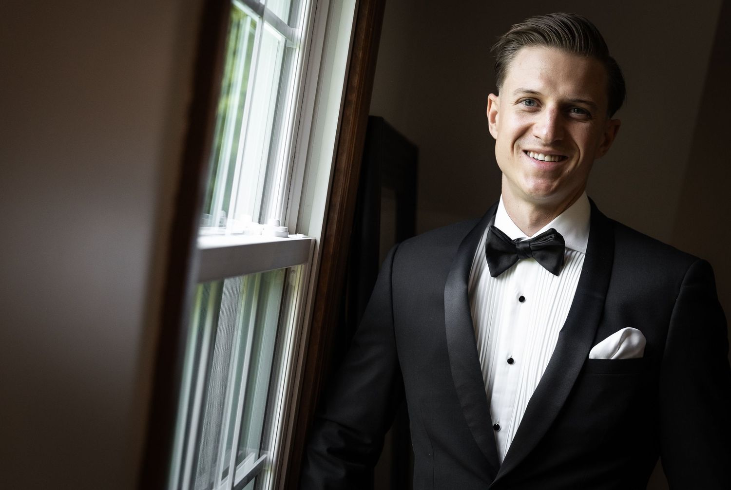 Classic formal portrait of groom in tuxedo by window.