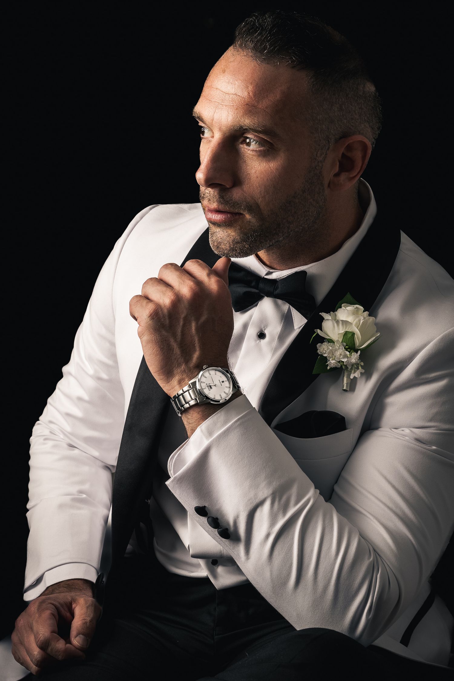 Groom in white tuxedo and black bowtie poses thoughtfully against dark background.