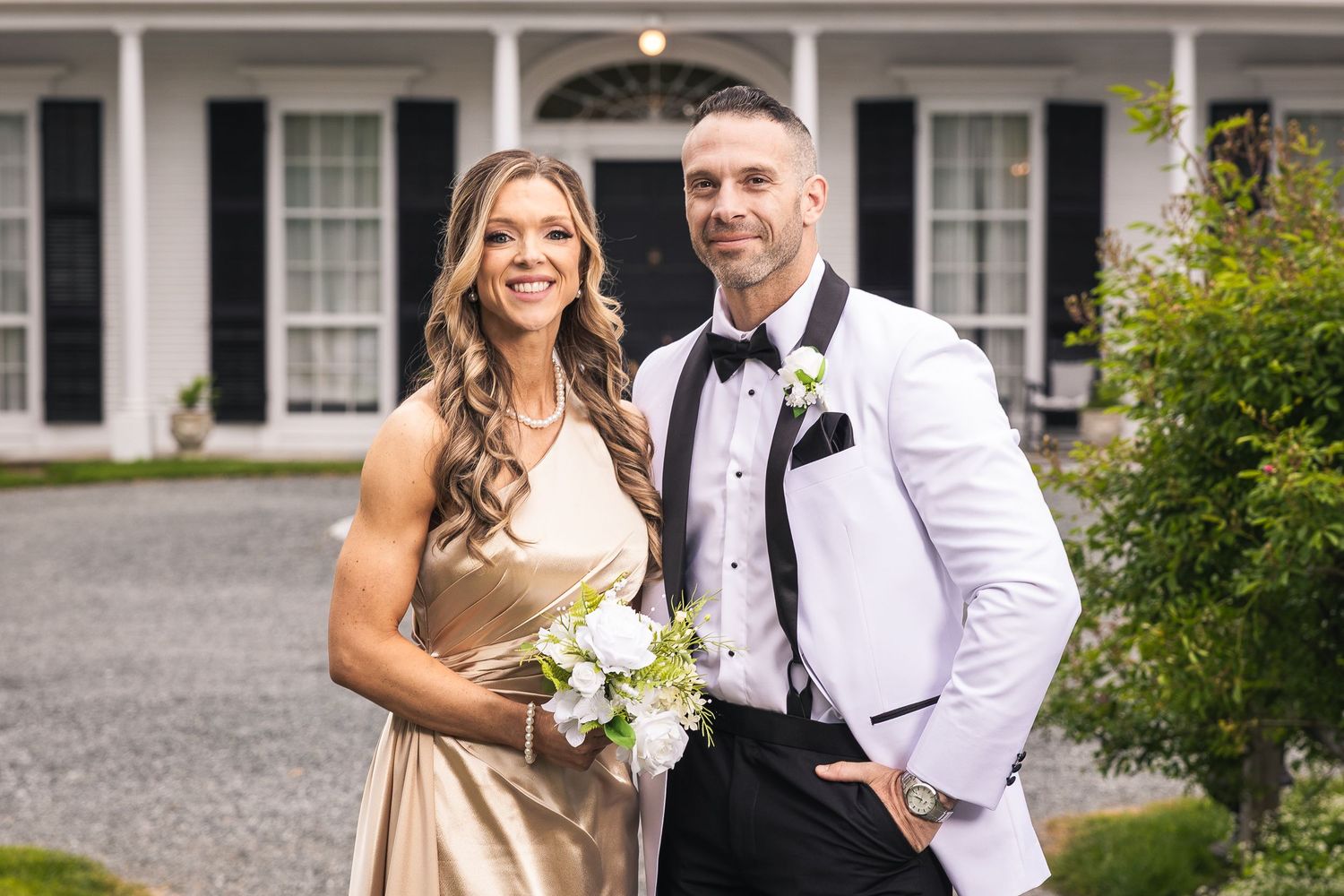 Wedding couple poses in front of a grand white colonial style mansion with black shutters.